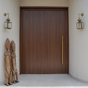 Projetti — Une porte en bois sombre et lisse, entourée de murs blancs, avec deux lanternes murales dorées de chaque côté. À gauche, deux sculptures en