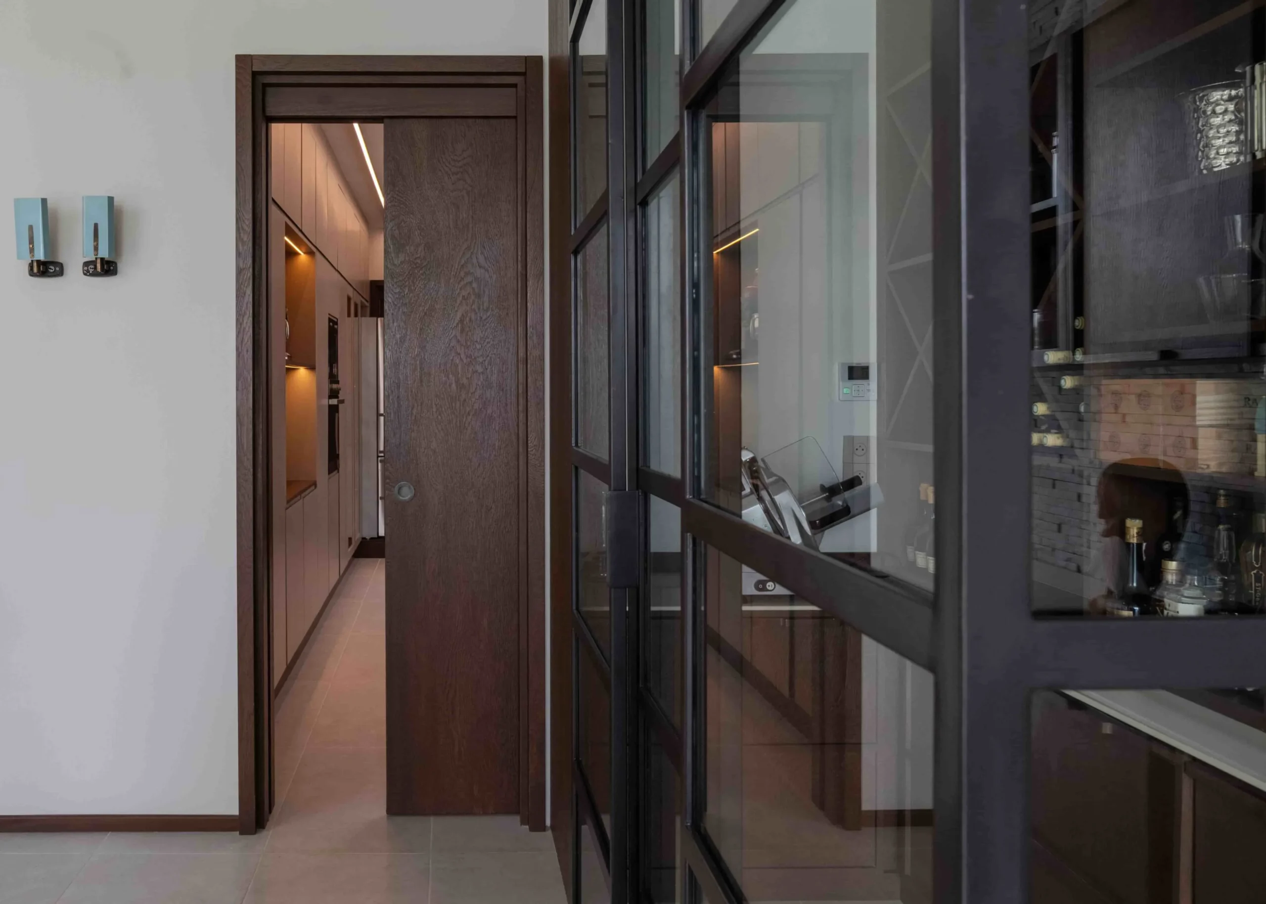 Projetti — Une porte en bois sombre s'ouvre sur un couloir éclairé, donnant accès à un salon contemporain avec une grande baie vitrée reflétant un bar