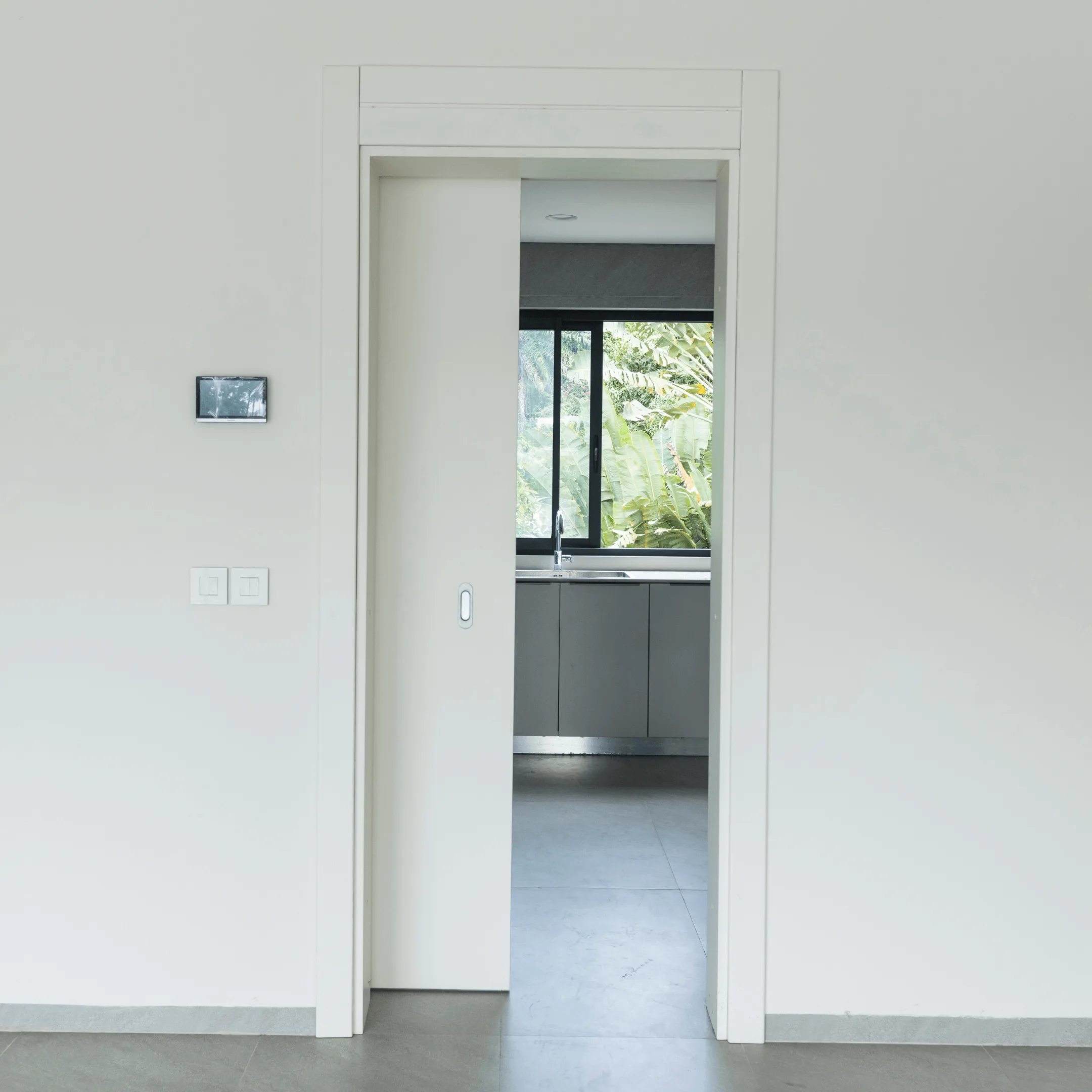Projetti — Salle de bain moderne et lumineuse Projetti — Une porte entrouverte donne accès à une salle de bain épurée aux murs blancs, avec une vasque encastrée sous une fenêtre laissant entrer un