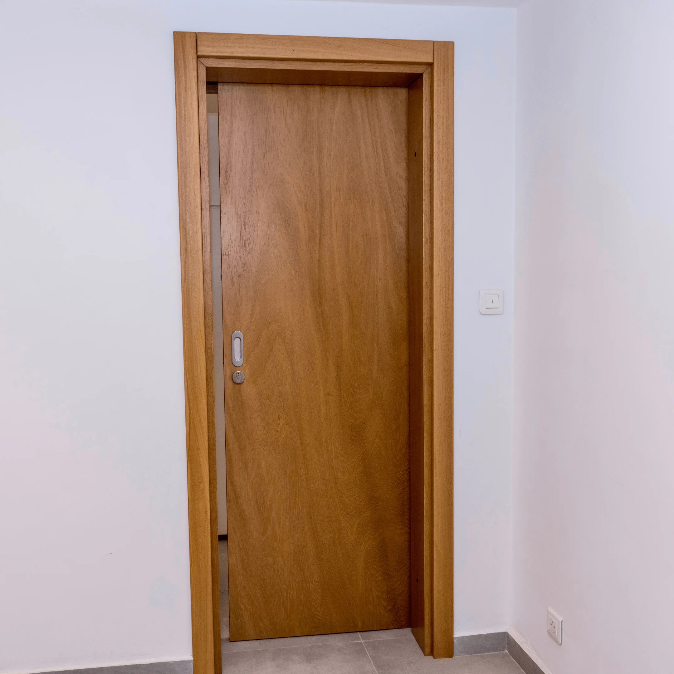 Projetti — Porte intérieure en bois clair Projetti — Une porte intérieure en bois clair, avec un cadre en bois naturel et une poignée ronde argentée, située dans un couloir blanc avec un sol c