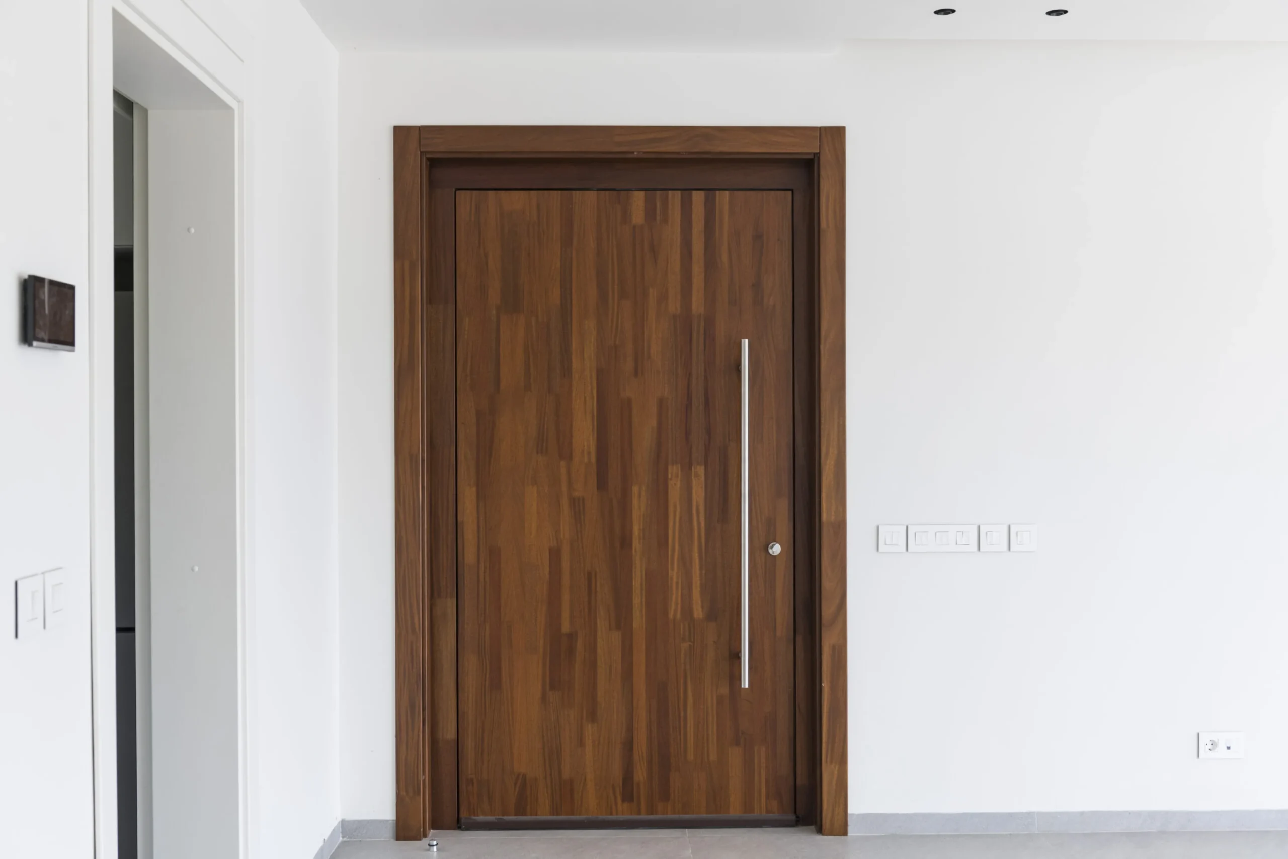 Projetti — Une porte d'entrée en bois massif aux veinages visibles, montée sur un cadre sombre, avec une poignée verticale argentée et un mur blanc ép