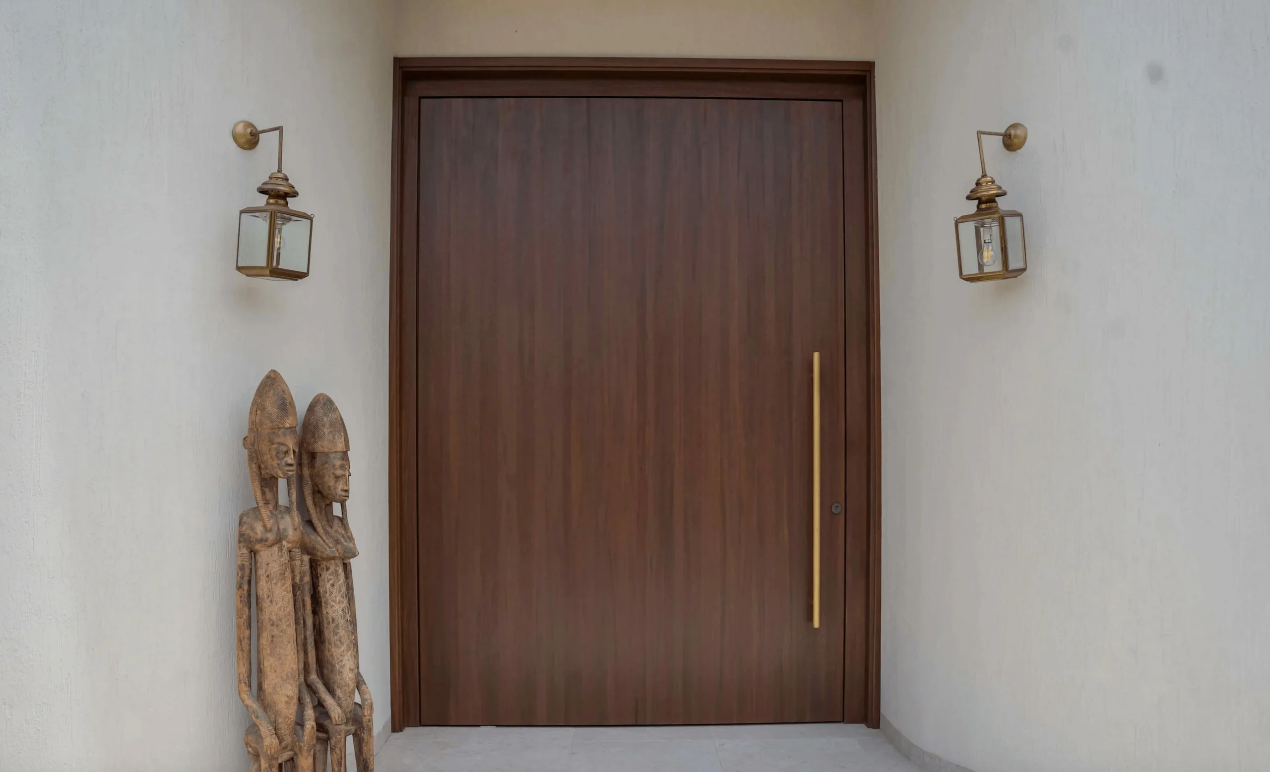 Projetti — Une porte en bois sombre et lisse encadrée par deux appliques murales en métal doré, avec deux sculptures africaines stylisées en bois plac